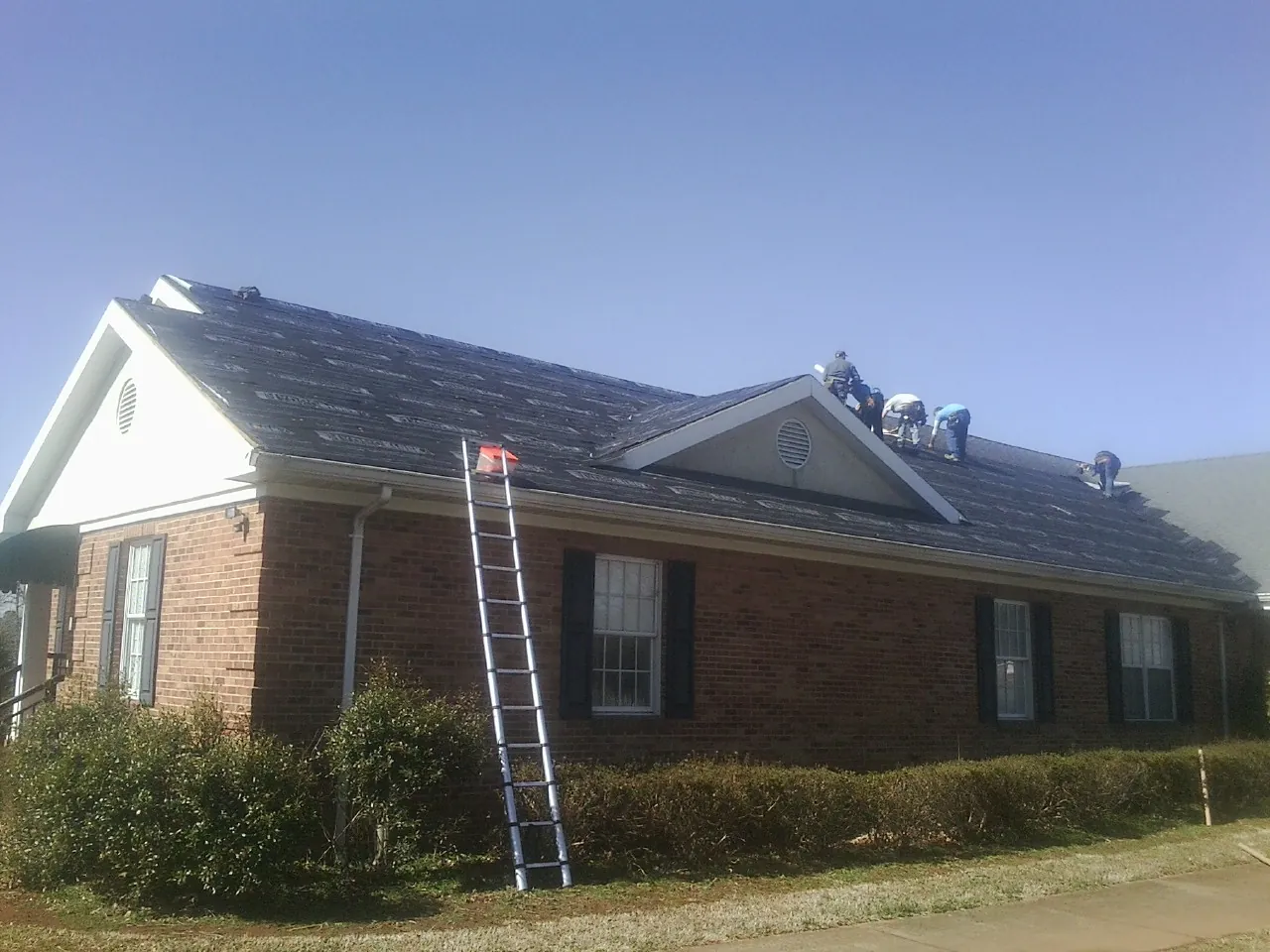 Crew tearing shingles off a brick ranch roof with a ladder set — Emergency Roof Repair work by our crew in Winterville