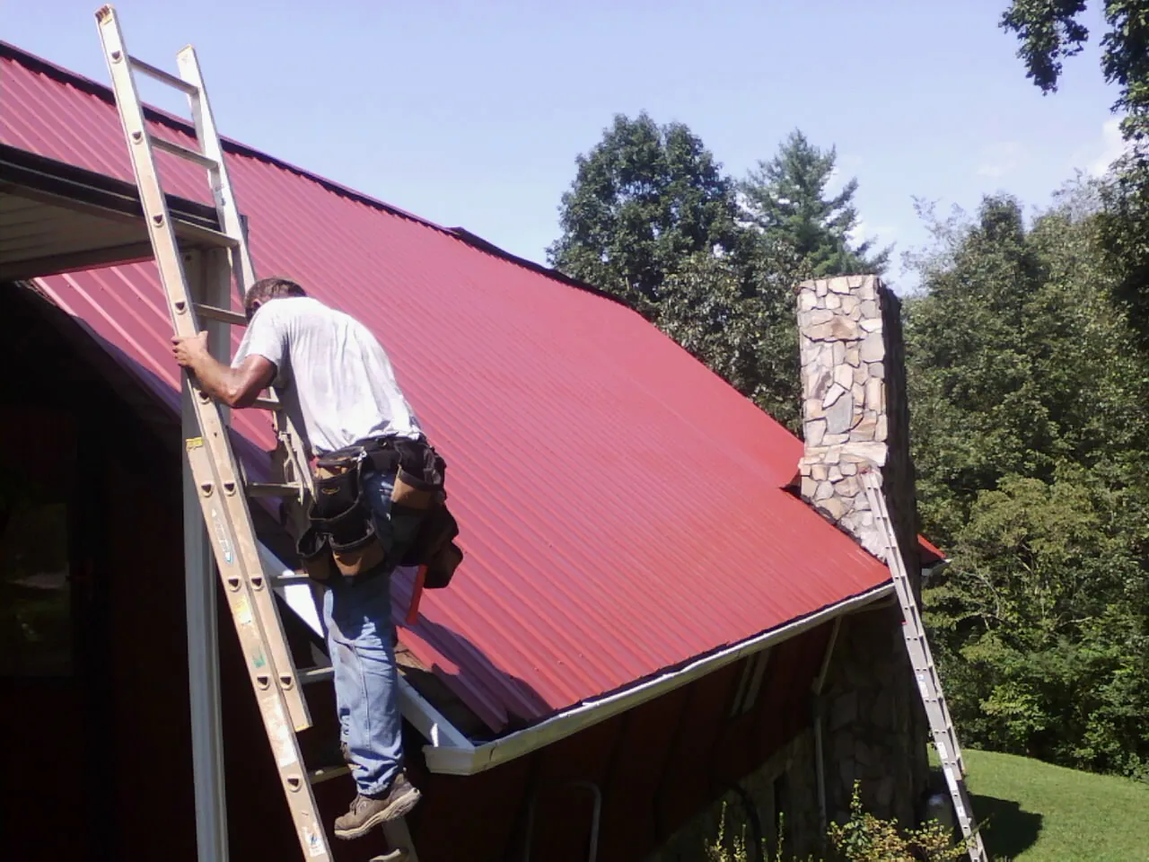 Roofer on a ladder beside a red metal roof and stone chimney — Soffit & Fascia Repair by Roofing Brothers in Winterville